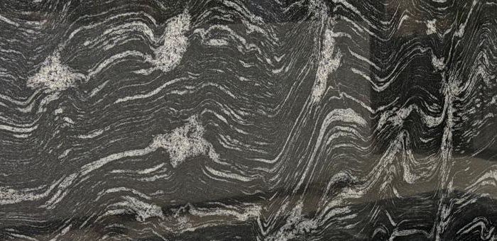Black granite slab with sweeping white and silver wave-like veining, standing in the Deepak Granito yard