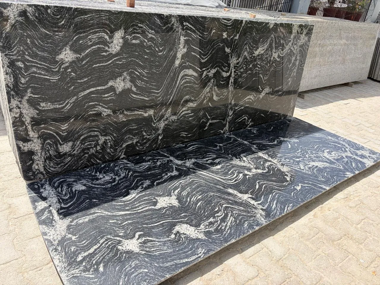 Black granite slab with sweeping white and silver wave-like veining, standing in the Deepak Granito yard